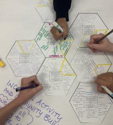 Multiple students' hands are writing and adding to a hexagon note.
